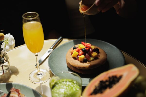 A person pours syrup over a pancake topped with diced mango, watermelon, and blueberries. Nearby are a glass of orange juice, a green smoothie, and a halved papaya on a table.