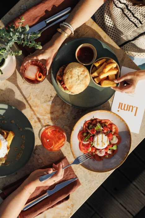 Overhead view of a table with a sandwich, chips, salad, drinks, and hands reaching for food. A menu with “lumi” is visible on the marble tabletop.