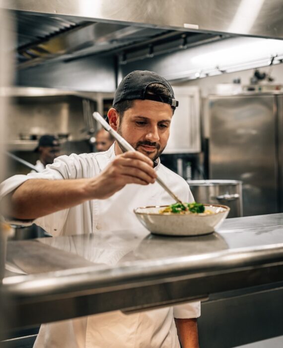 A chef in a white uniform and black cap carefully garnishes a dish in a commercial kitchen, using a spoon to add detail, with stainless steel appliances and another staff member visible in the background.