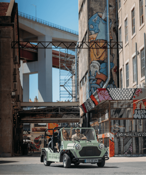 A small green open-top car drives through an urban street with two people inside. Colourful graffiti covers nearby buildings, and a large bridge is visible in the background.