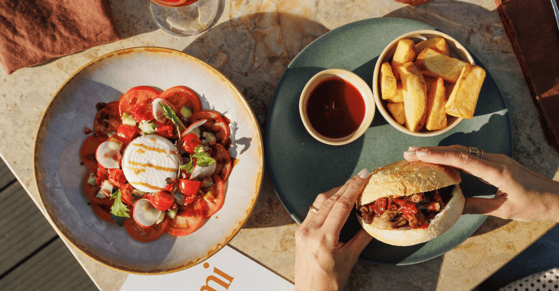 A person holds a burger above a plate with a side of potato wedges and dipping sauce, next to a bowl of tomato and burrata salad.