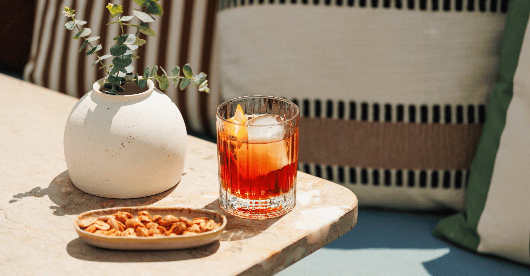 A rocks glass containing a dark amber cocktail with a large ice cube and an orange twist, set on a marble table next to a small dish of peanuts and a vase with eucalyptus.