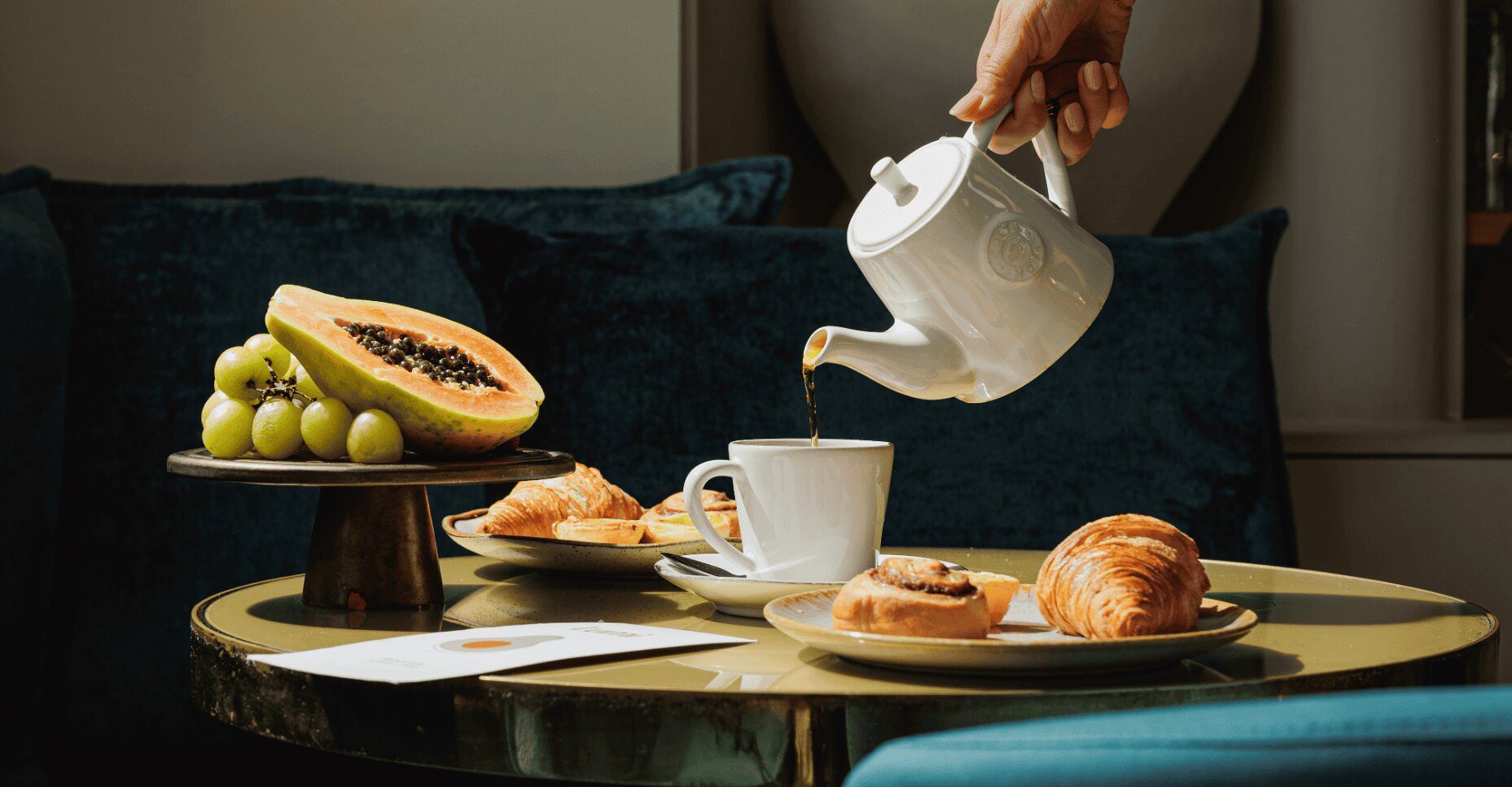 A hand pours tea from a white teapot into a cup on a table set with pawpaw, grapes, and pastries.