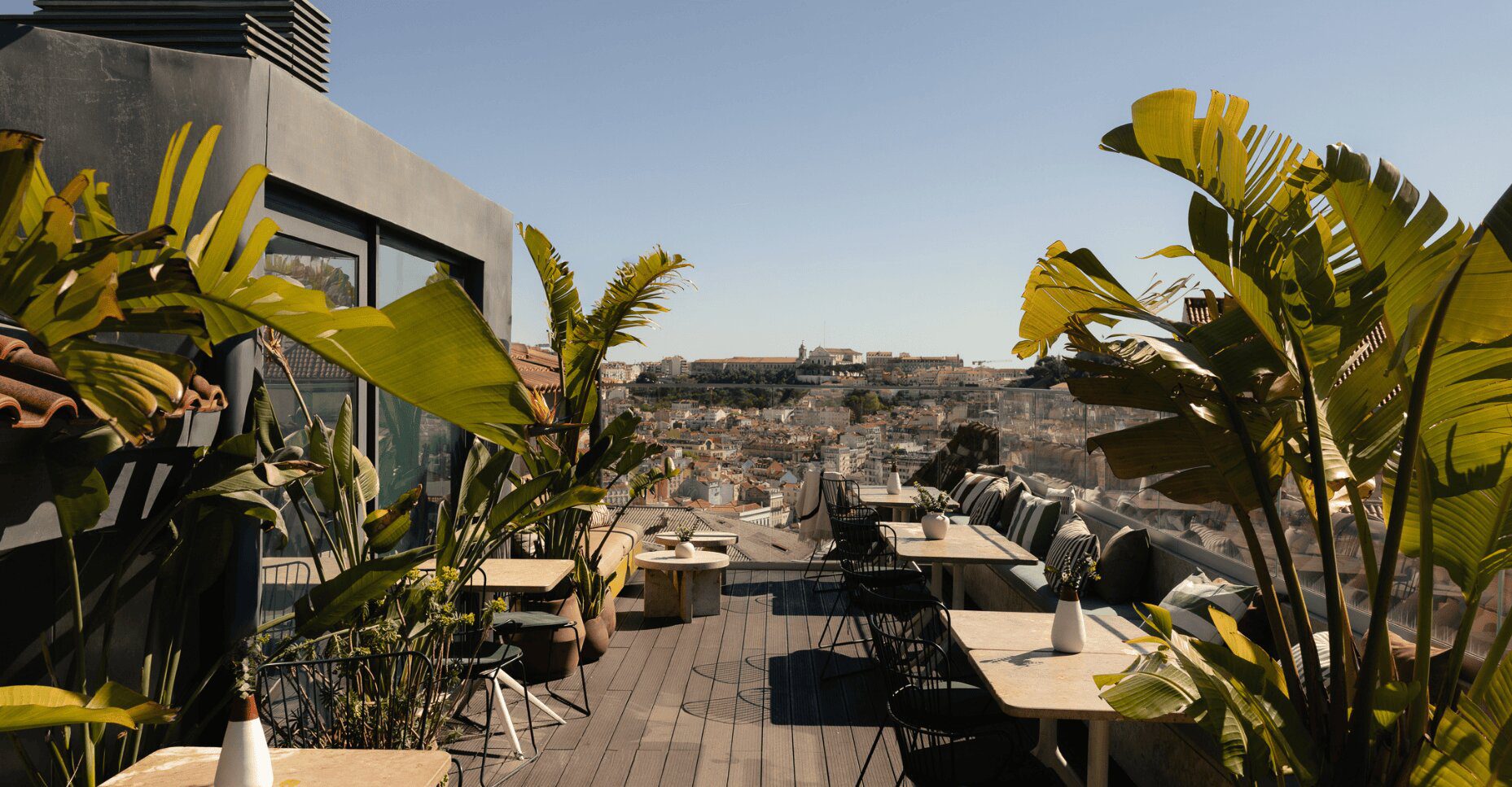 Rooftop terrace seating with dining tables, chairs, and potted plants overlooking a city skyline.