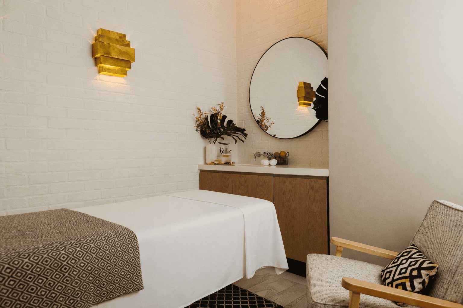 A spa treatment room featuring a massage table, a small dressing table with a round mirror, and a wooden armchair.